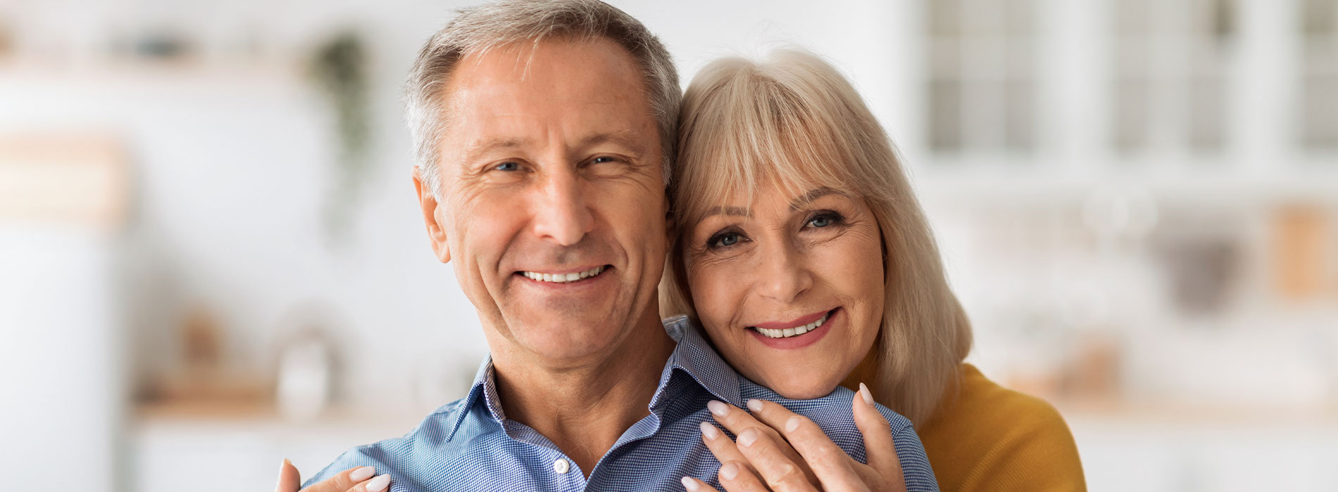 The image shows an older man and woman, possibly grandparents, embracing each other with smiles on their faces, set against a blurred domestic background.