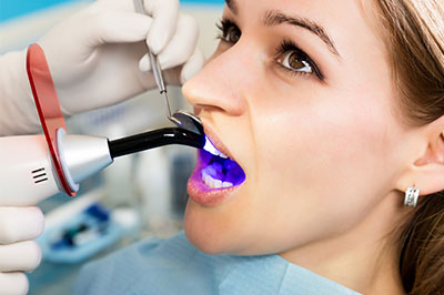 The image shows a woman sitting in a dental chair with a device attached to her mouth, likely for teeth whitening or cleaning, while a dental professional is using a machine with a blue light on her face.