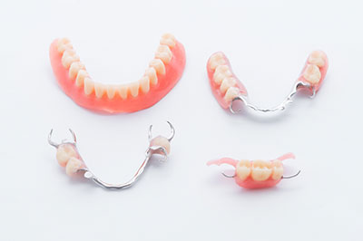 The image shows a set of dentures with a partial upper plate featuring an artificial tooth on a white background, placed next to a silver wire frame with a toothpick inserted into one side.