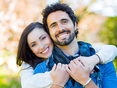 A man and woman are embracing each other in a joyful pose  they appear to be young adults with smiles, standing outdoors on what seems to be a pleasant day.