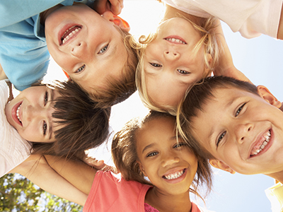 A group of children with different expressions, smiling and laughing, posing together outdoors under a bright sky.