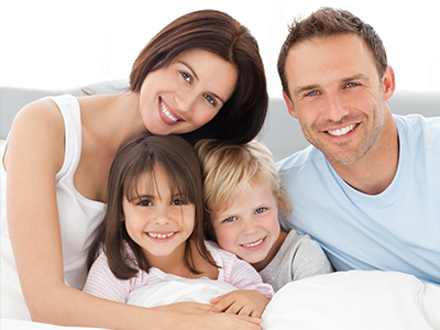 The image features a family of four - a man, a woman, and two children, all smiling and posing together on a bed.