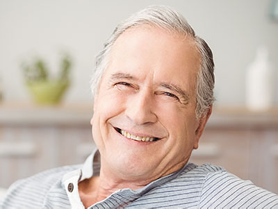 The image shows a smiling older man with gray hair, wearing a blue shirt, seated comfortably in a chair indoors, looking directly at the camera.