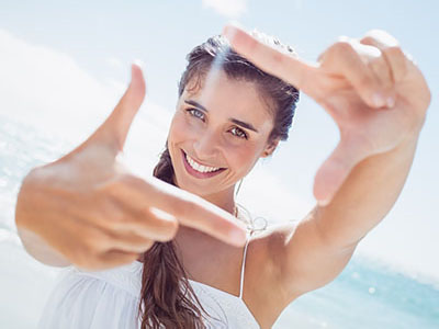 A woman with long hair smiles and holds up her hand forming a peace sign against a bright background.